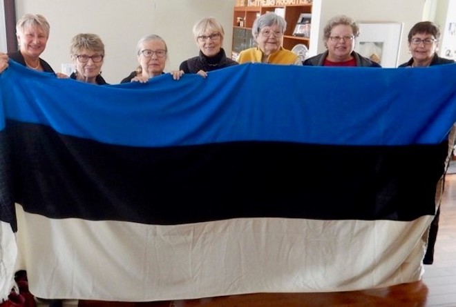 Hoides õrnalt Pika Hermanni tornis lehvinud sini-must-valget lippu, TES juubelikomitee liikmed tervitavad EV100 juubelipidustuste eel: vas Elle Rosenberg, Ellen Leivat, Anne-Liis Keelmann, Maaja Matsoo, Anne Orunuk, Epp Aruja, Helle Arro.