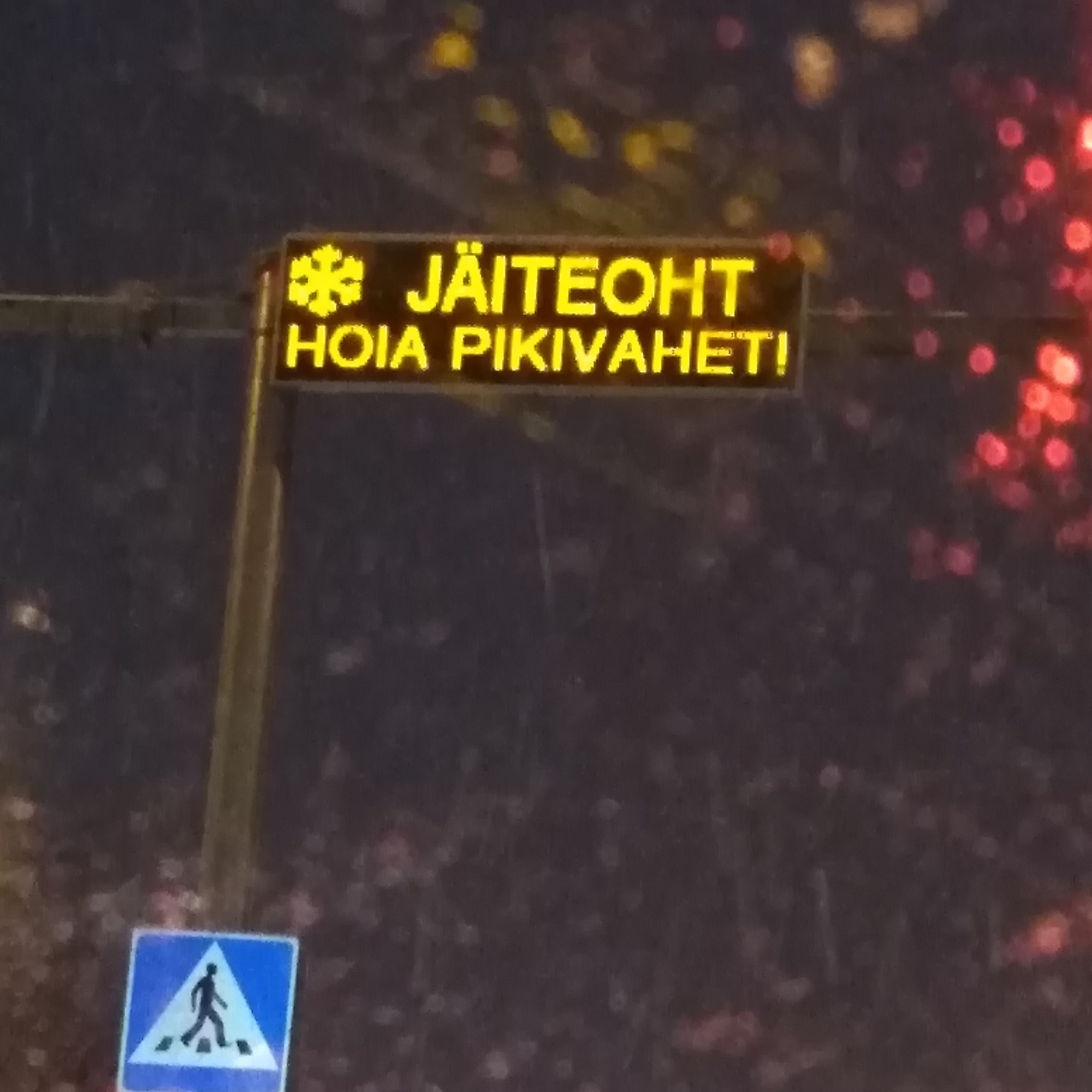 When the temperature is around null (zero, the freezing mark, 32°F), this sign above Sõle tänav in Põhja-Tallinn lights up. Photographed through an icy tuule/klaas (windshield) while waiting punase tule taga (at a red light), it was mounted this past fall above the valgus/foorid (traffic lights) and jala/käijate üle/käigu/rada (pedestrian crosswalk) where crowds of kids cross to and from Pelgu/linna Gümnaasium (High School). Foto: by Riina Kindlam (2019)