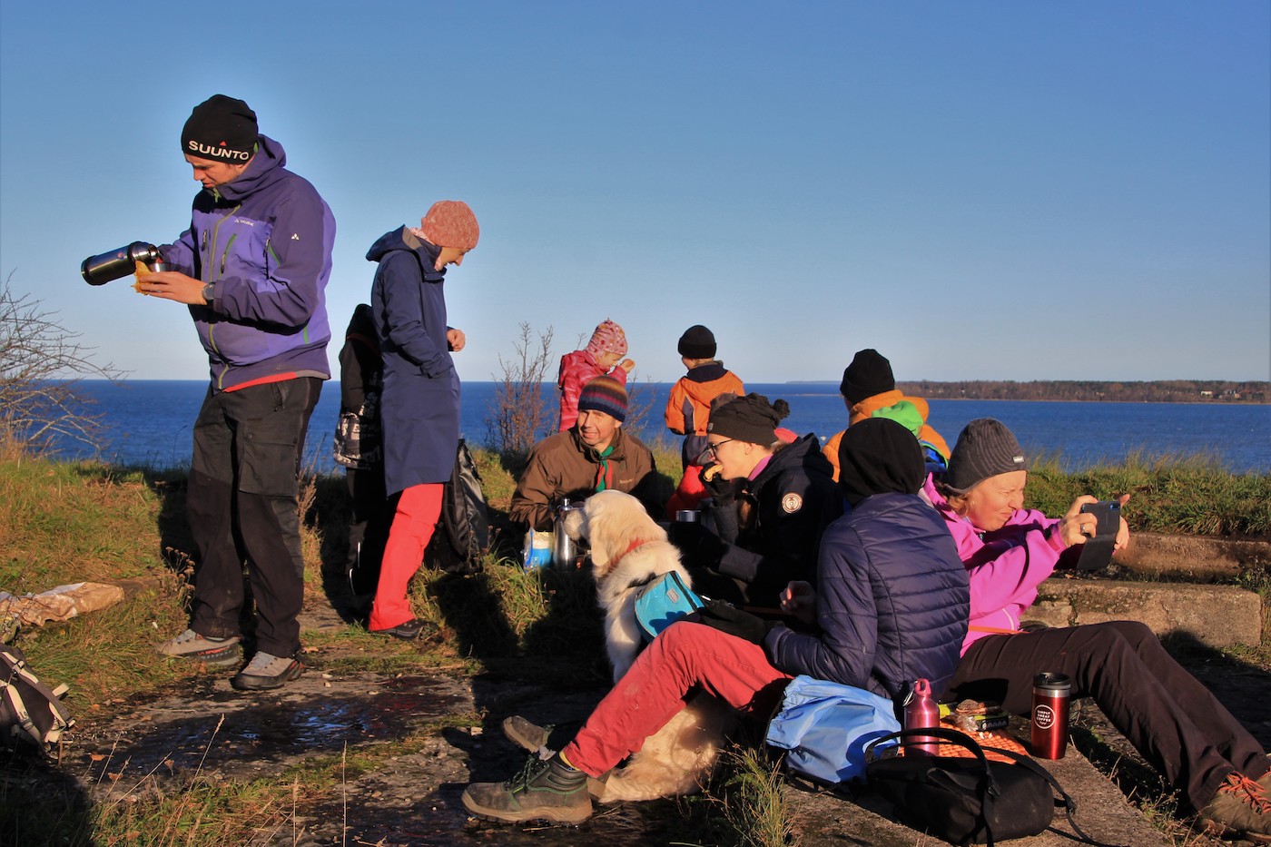 Pangalt ehk panga pealselt laskuti panga alla veepiirini (küll mitte köiega vaid jalgsi) ja siis roniti taas üles. Foto ja matkaseltskond oli väga kirev. Paberlehelugeja kahjuks ei näe, kui rukkilillesinine on meri ja kui punased paari matkaja püksid. Koer Wanda keset fotot on see eest elevandiluukarva. Ja jalge all on paas. Taamal paistab Kakumäe poolsaar. On varajane pärastlõuna, kuid päike paitab juba vägagi madalalt. Foto: Nele-Kai Loorits (2020)