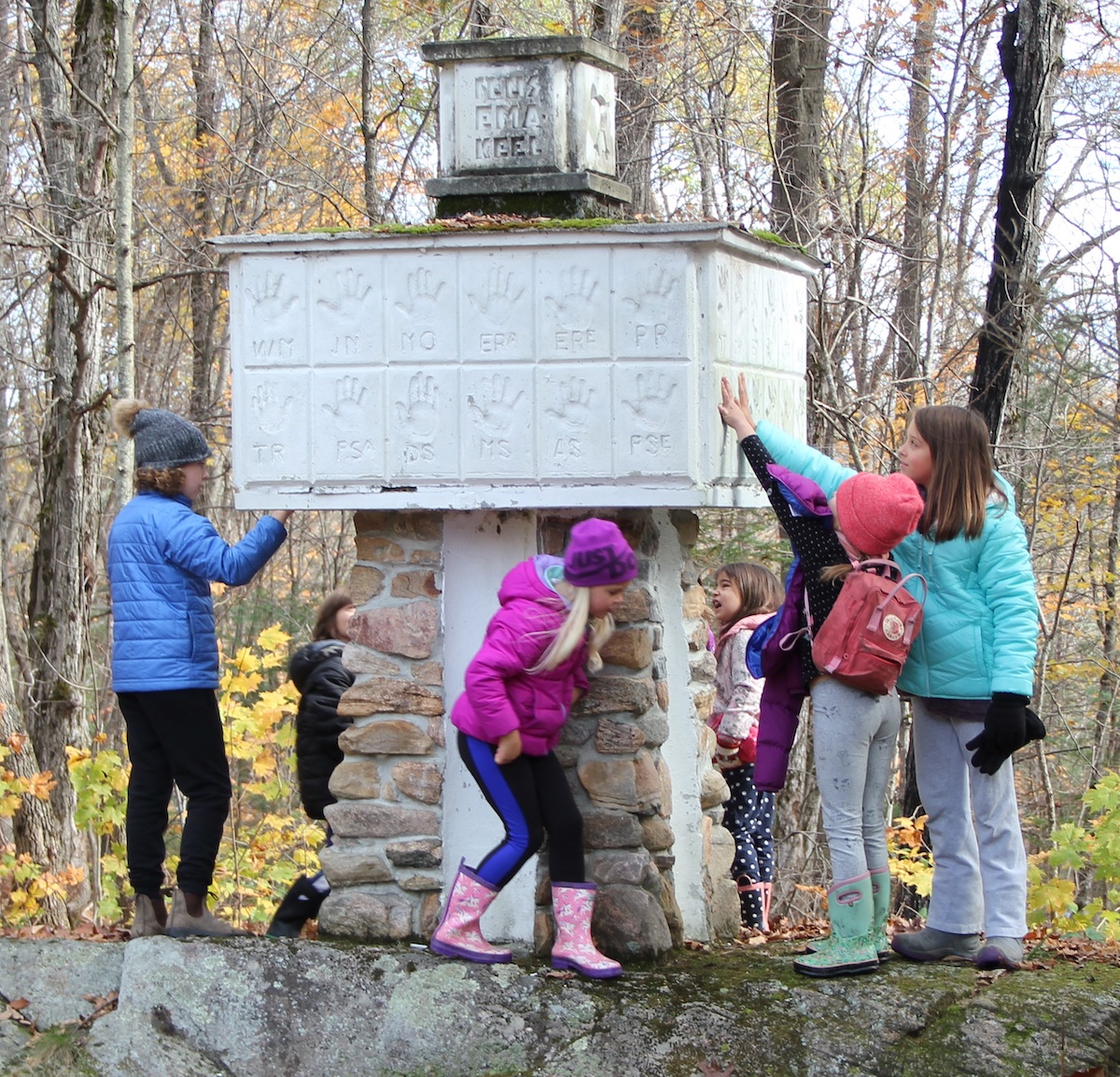 ILUSA EMA/KEELE SAMMAS on au/sammas ehk monument eesti keelele. See püstitati Põhja/naela (*Polaris, the North Star*) suur/laagris 33 aastat tagasi! Hellakesed ja hundud kogusid selle jaoks kive ja juhid koos vana/skautidega (isad ja vana/isad) ehitasid samba. Oma käe/jälje pani vedela betooni (*concrete*) sisse 48 noort, andes lubaduse (tõotuse, *pledge*), et nad IIAL EI UNUSTAKS OMA EMA- või ISA/KEELT – EESTI KEELT. Iga käe/jälje juures on noore nime/tähed, kelle käsi see on. Kas Marika (vasakul), kes oli esimest korda Kotkajärvel, oskas arvata, et tema isa, Erik Lepiku käe/jälg on seal? Kas Helena (jalutab samba alt läbi), leidis oma isa Andres Kolga käe/jälje? Metsas töödega abistava Markuse isa Eerik Randsalu käe lubadus on ka sambal. Ja mina, kes ma teile kirjutan, kuna olin siis Lembitu Maleva hundu/juht. Kõiki seekord ühe foto peale ei saanud, aga pildile on jäänud käsi sambale sirutavad Mia-Liina Hess ja Siena (Vahi) Franzoi. Vahelt paistab täpiliste pükstega Silvi (Voksepp) DiBiase. Õnneks oma jälje eesti keele püsimisse – kiidu/väärt keelega pusimisse – saad panna ka ilma kätt tsementi vajutamata. Seda annab iga kell mõtteliselt teha. JÕUDU! Foto: Maimu Mölder (2021)