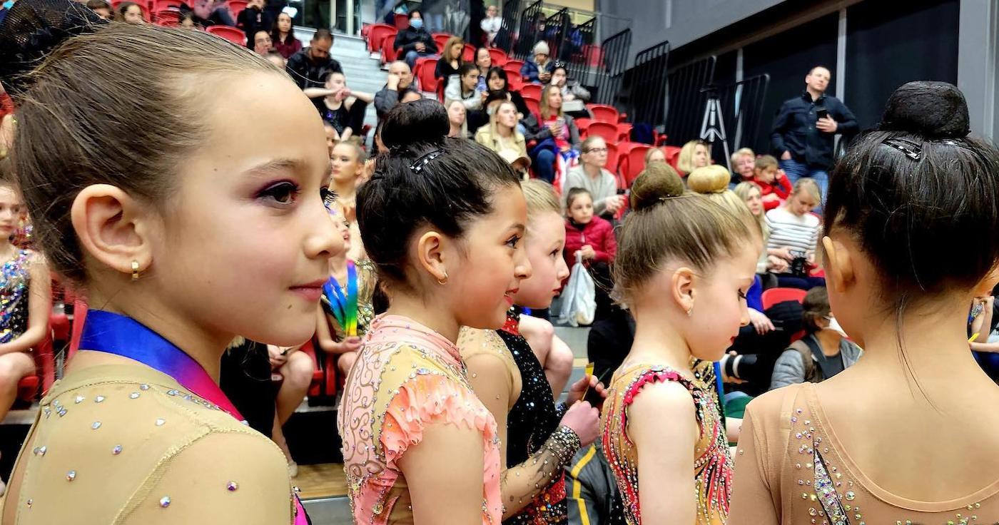 Young gymnasts during their awards ceremony with members of the audience visible in the background. Photo by Shelley Jacklin