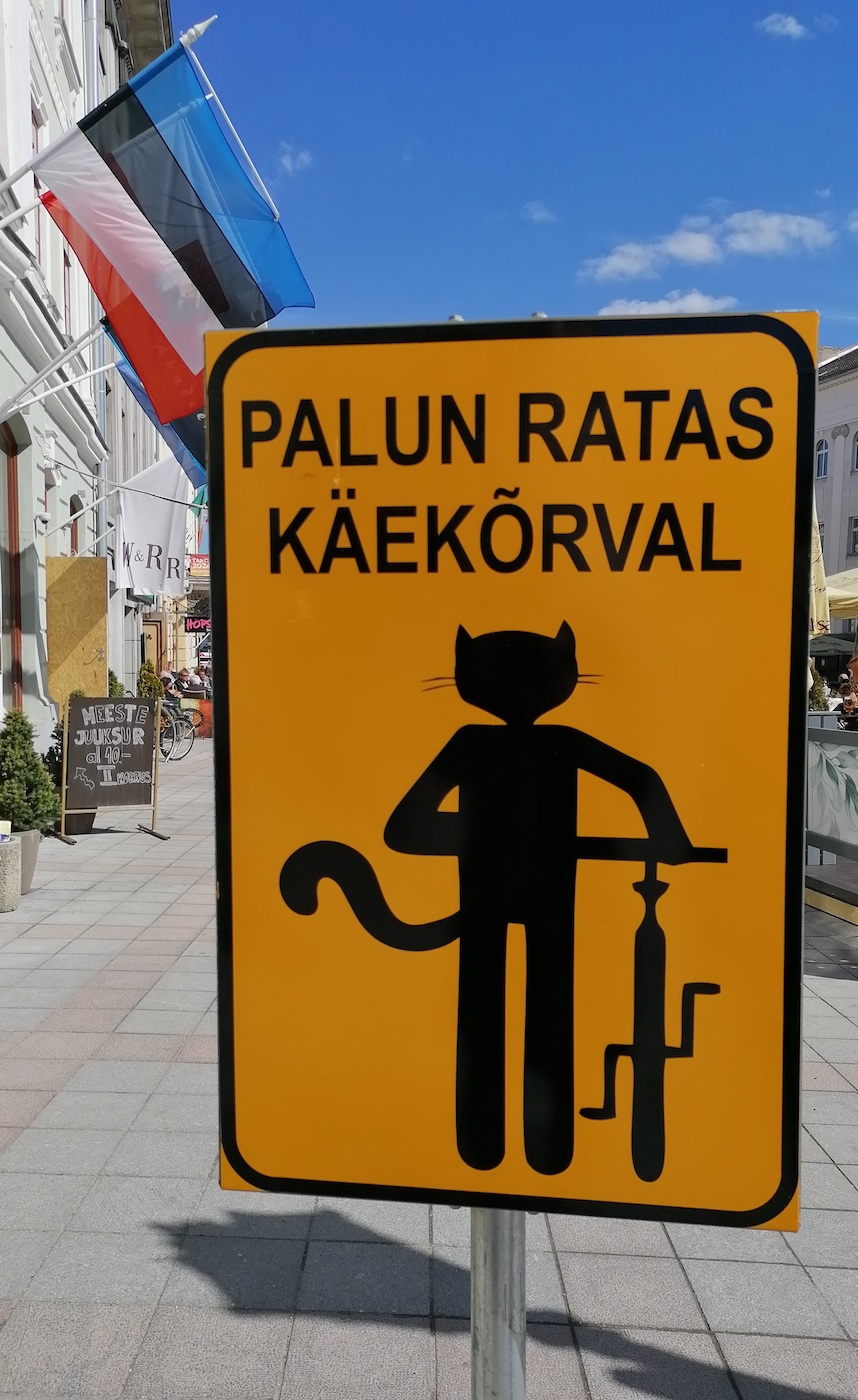 Tartu Rae/koja platsil antakse teada, et platsil peavad kõik, ka kassid, tulema JALGratta sadulast (*saddle*) maha ja kõndima ratast käe/kõrval lükates. Ilmselt kehtib see palve kõikidele teistele liiklejatele, kellel üks/kõik millised RATTAD all. Natuke maad eemal, Ema/jõe kaldal on sarnased kiisu/ratturiga (*kitty-cyclist*) sildid, kus palutakse olu/korrale vastavalt (*according to the situation*) käituda: ,,Kui rahvast on palju, palun võta ratas käe/kõrvale! Aitäh!“ Foto: Riina Kindlam