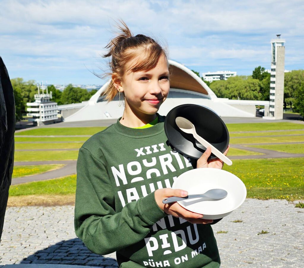 Noor koorilaulja korduvkasutatavate (reusable) toidunõudega. Tema taga paistab laululava ehk laulukaar ja torn, mille otsas sütib laulupeotuli. Foto laulu- ja tantsupeo Facebooki lehelt.