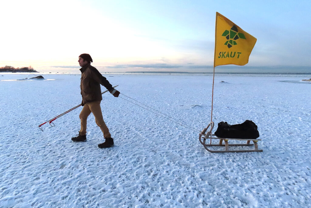 Maade/avastaja (explorer) Vaida küla Krattide hundu/juht Radames Haiba läheb JÄÄ/retkele 20. hundi/kuul loojangu (sunset) eel Põhja-Tallinnas Koplis. Skaudi/lipp paistab kaugele – oluline on olla nähtav. Kelgul on uisud, mis kohe jalga tõmmati, et nautida Paljas/saare lahe (bay) jääd. Ka läbi lume/kihi (kiht, layer) andis sõita. Kohati oli ka MÕNUSaid SILEdaid jää/laike (patches of ice). Fotost paremale paistsid Tallinna SADAMast (from the harbour) Helsingisse suunduvad parv/laevad (ferries). Foto: Riina Kindlam