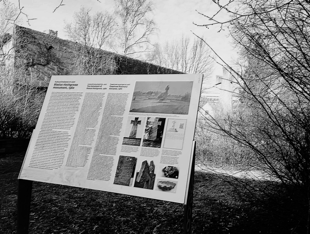 The information board near the Hochgreve Cross in Kitseküla, with the monument visible in the background through early spring branches (photo: Jerry Mercury)