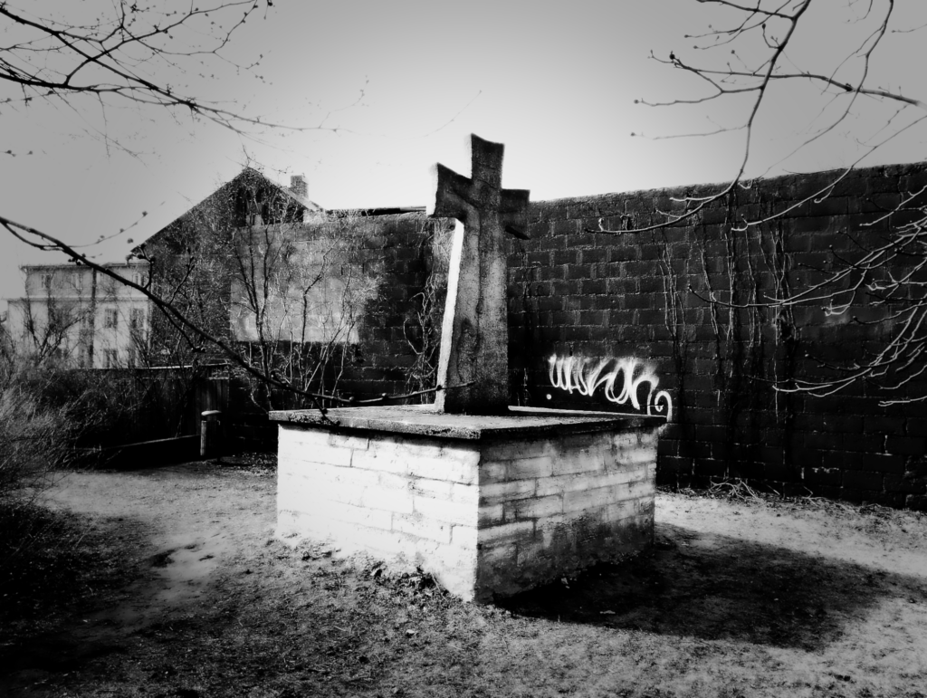 The Hochgreve cross in early spring, against a dark-painted brick wall with bare vines tracing its surface. A fresh graffiti tag has already appeared, and the creeping plants are half-covered in black paint.
Photo: Jerry Mercury