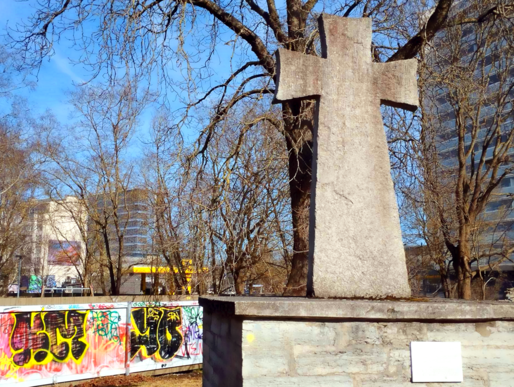The Hochgreve cross on a bright spring day. Behind it, a fence layered in graffiti, and beyond that, the yellow canopy of a gas station. Photo: Jerry Mercury